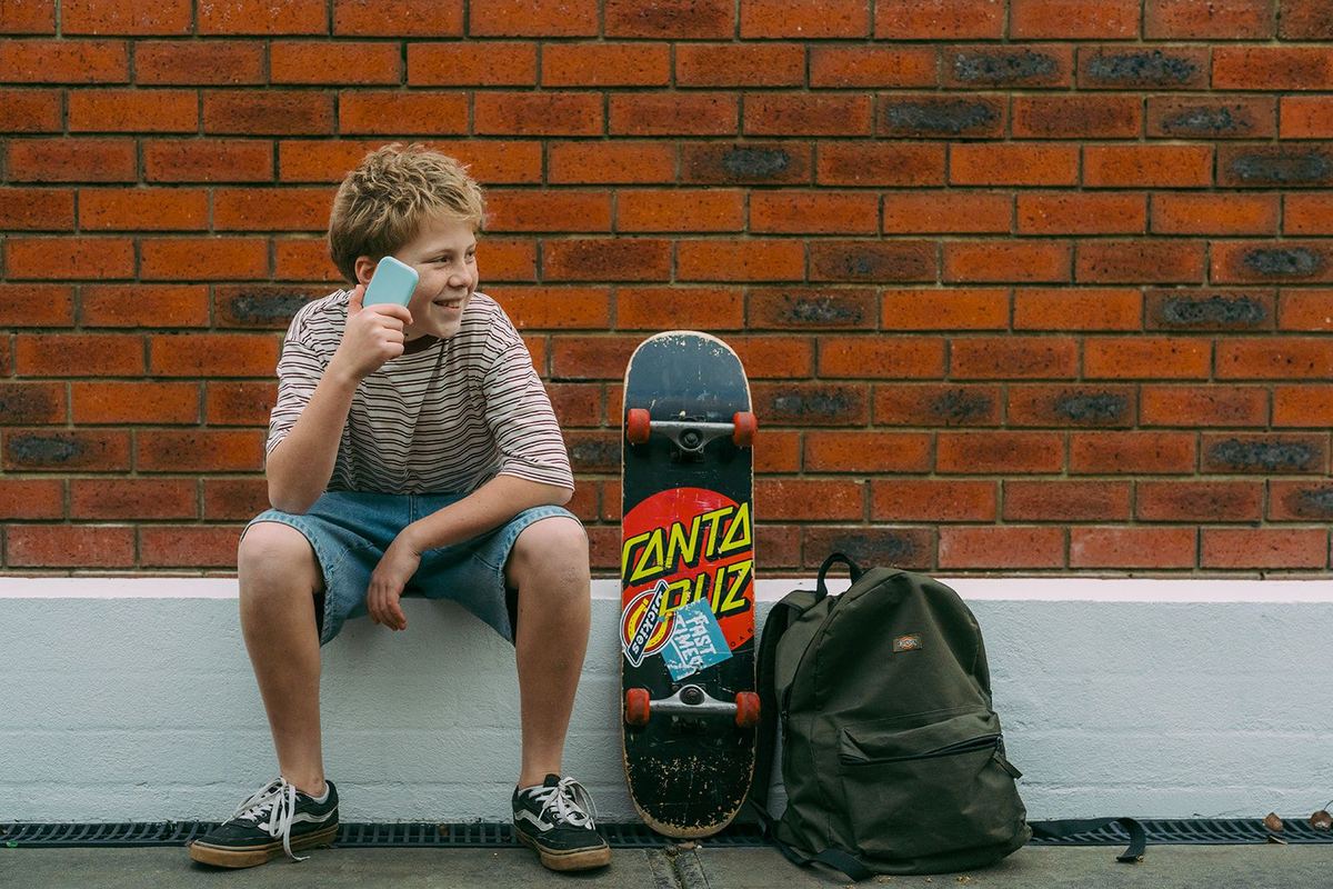 A boy sitting on a low wall with his skateboard and backpack, holding a Waffle Go phone to his ear and smiling.